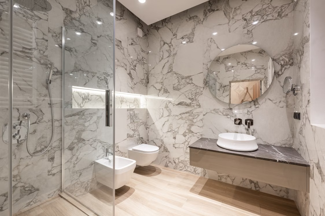 Baño reformado con revestimiento de mármol, ducha de cristal y mueble suspendido